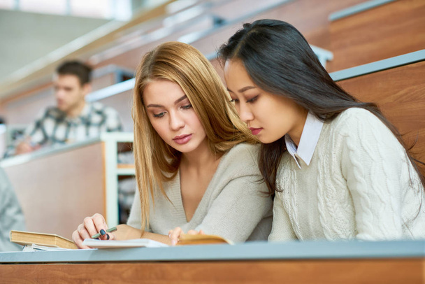 Two beautiful girls reading notes sitting at desk in lecture hall of modern college, copy space - Foto, imagen