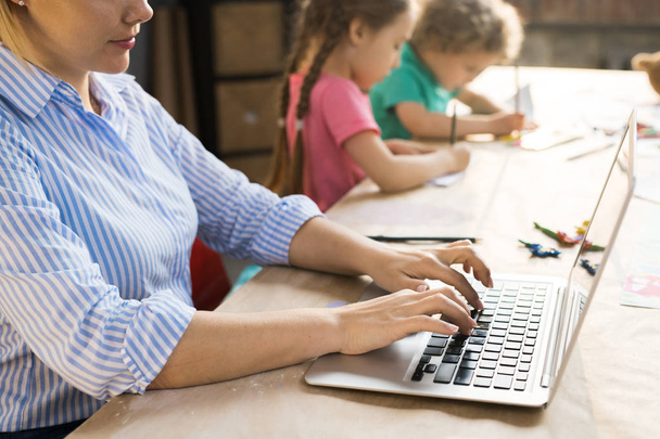 Vista lateral de la joven madre sentada y escribiendo en el portátil mientras sus hijos dibujan en la mesa
 - Foto, Imagen