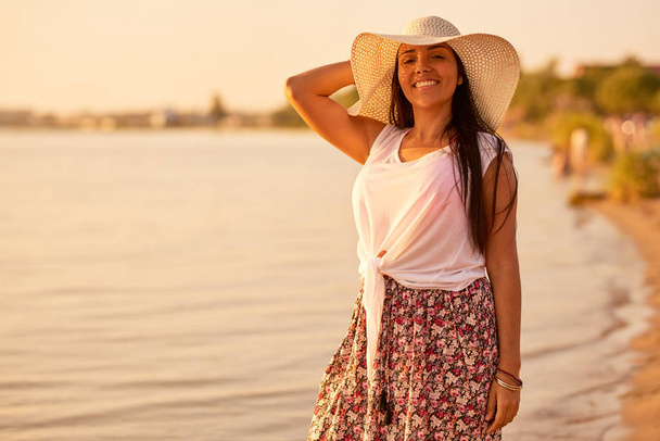Lachende vrolijke mooie Spaanse vrouw in zomer outfit aanpassen zon hoed en camera kijken tijdens het staan op strand - Foto, afbeelding