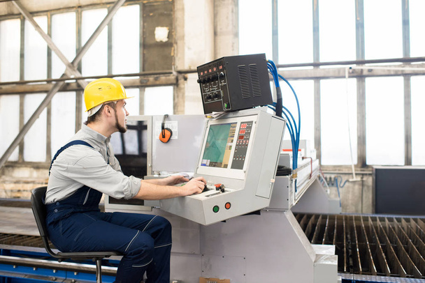 Serious concentrated skilled young manufacturing specialist in hardhat sitting at computer of manufacturing machine and choosing setups for conveyor belt in factory shop - 写真・画像