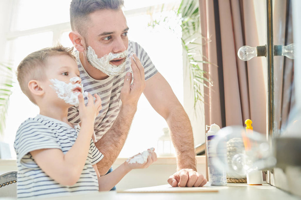Handsome young man and his little son with shaving foam on their faces having fun together at spacious bathroom with panoramic window - 写真・画像