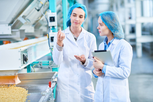 Портрет двух молодых работниц в лабораторных халатах, стоящих на конвейере с макаронами в цехе чистого производства
 - Фото, изображение