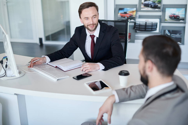 Retrato del gerente de ventas guapo barbudo hablando con el cliente mientras está sentado en el escritorio en la sala de exposición de automóviles
 - Foto, imagen