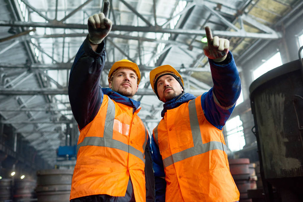 Group portrait of confident bearded workers wearing hardhats and reflective vests pointing at something with index fingers while standing at production department of modern plant - Photo, Image