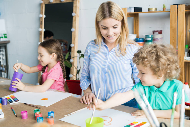 Sorridente giovane insegnante in studio d'arte aiutare i bambini nella pittura
 - Foto, immagini