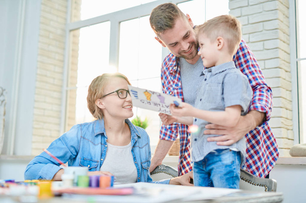 Portrait of modern young family with cute little son smiling happily looking at each other while  celebrating Fathers day together  standing against big windows at home. - Photo, Image