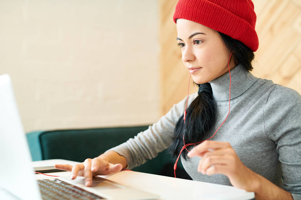 Retrato da jovem mulher moderna usando chapéu de gorro usando laptop enquanto faz trabalho freelance no café e ouvindo música
 - Foto, Imagem