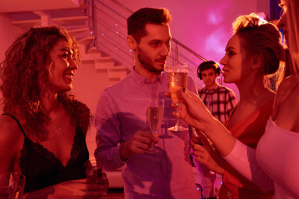 Стривайте портрет красивого чоловіка в оточенні красивих молодих жінок і спілкуйтеся щасливо, п'ючи шампанське під час домашньої вечірки в червоному світлі
 - Фото, зображення