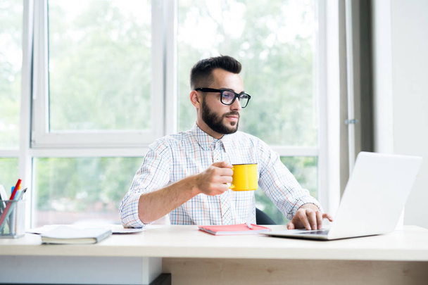Retrato del hombre de negocios barbudo de moda que trabaja en el escritorio en la oficina usando el ordenador portátil y disfrutando de una taza de café, espacio para copiar
 - Foto, imagen