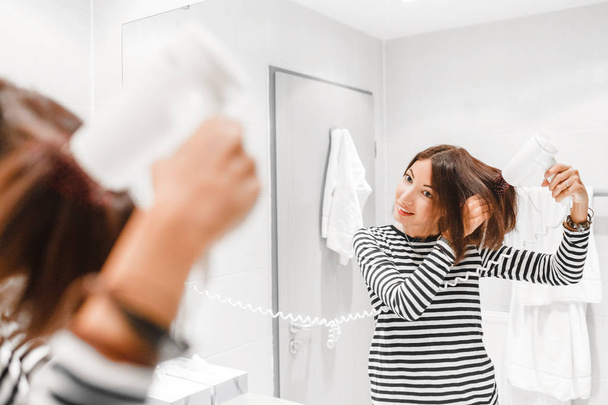 Giovane donna felice sorridente e asciugandosi i capelli con asciugacapelli vicino allo specchio in bagno
 - Foto, immagini
