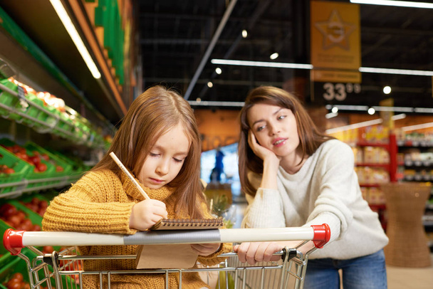 Портрет молодой женщины, покупающей продукты с дочерью в супермаркете, читающей список покупок и сидящей в корзине
 - Фото, изображение