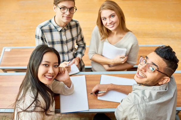 Vista de ángulo alto en el grupo multiétnico de estudiantes alegres mirando a la cámara y sonriendo sentado en el escritorio en la sala de conferencias de la universidad moderna
 - Foto, Imagen