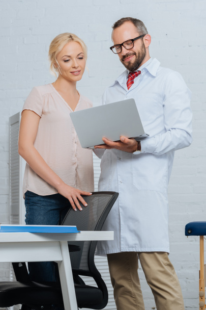 fisioterapeuta sorridente em casaco branco e paciente do sexo feminino usando laptop no hospital
 - Foto, Imagem
