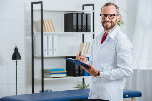 portrait de chiropraticien masculin souriant en manteau blanc et lunettes avec bloc-notes à l'hôpital
 - Photo, image