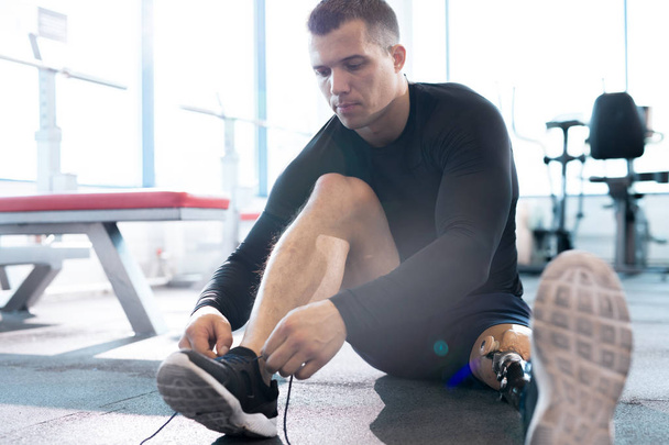 Retrato de comprimento total de desportista muscular com prótese perna amarrando sapato sentado no chão no ginásio moderno
 - Foto, Imagem