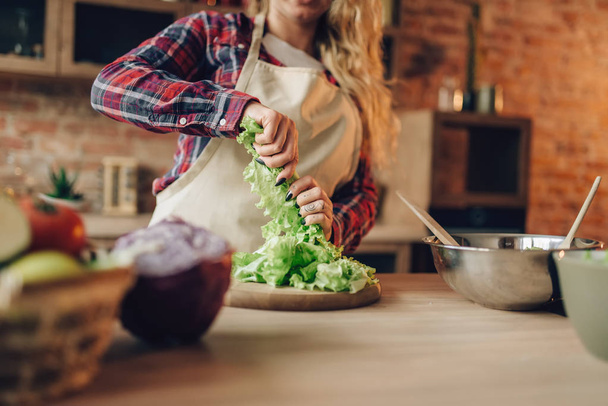 Νοικοκυρά στην ποδιά ετοιμάζει σαλάτα, κουζίνα, εσωτερικό σε φόντο. Μαγείρισσα κάνοντας υγιείς χορτοφάγους τροφίμων, προετοιμασία λαχανικών - Φωτογραφία, εικόνα