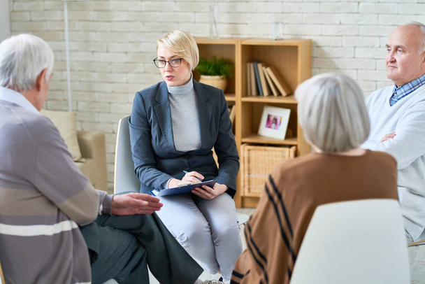 Retrato de psiquiatra loira vestindo óculos levando sessão de terapia de grupo para pessoas idosas em casa de repouso, espaço de cópia
 - Foto, Imagem