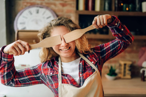 Playful housewife in an apron covers her eyes with wooden spatulas, kitchen interior on background. Happy female cook making healthy vegetarian food, salad cooking - Photo, Image