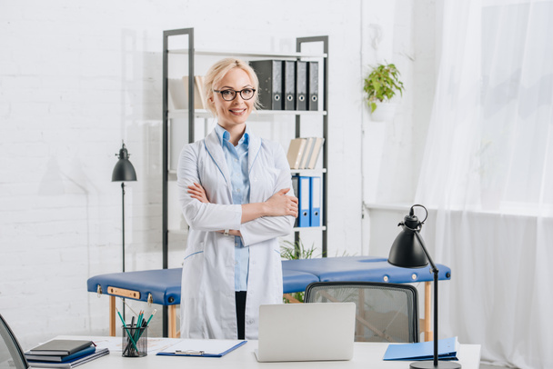портрет улыбающегося мануального терапевта в очках и белом халате, стоящего на рабочем месте в клинике
 - Фото, изображение