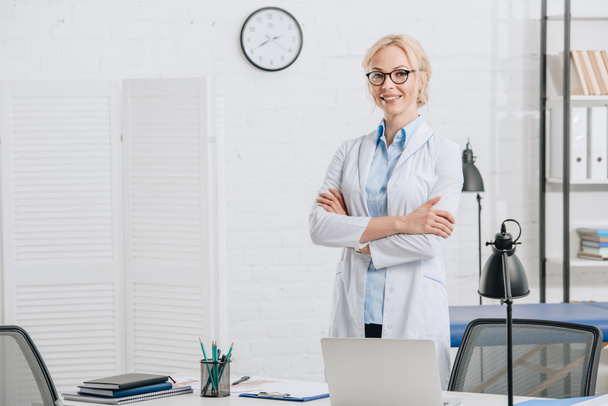 portrait of smiling chiropractor in eyeglasses and white coat standing at workplace in clinic - Photo, Image