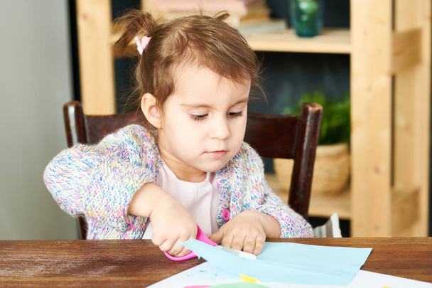 Portré lövés imádnivaló kicsi lány ül a fából készült asztal és ollóval, miközben üdvözlés kártya Mother's Day  - Fotó, kép