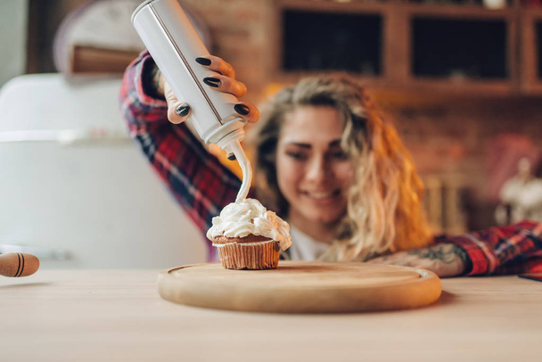 Housewife in apron covers sweet dessert with cream, kitchen interior on background. Female cook prepares fresh homemade cake. Domestic pie preparation - Photo, Image
