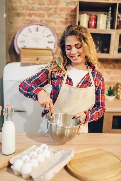 Lächelnde Hausfrau in Schürze, Teig in Schüssel, Kücheneinrichtung im Hintergrund. Köchin bereitet frischen hausgemachten Kuchen zu. Zubereitung von Kuchen - Foto, Bild