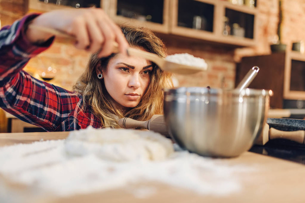 Young housewife in an apron puts flour in a bowl, kitchen interior on background. Female cook prepares fresh homemade cake. Domestic pie preparation - Photo, Image