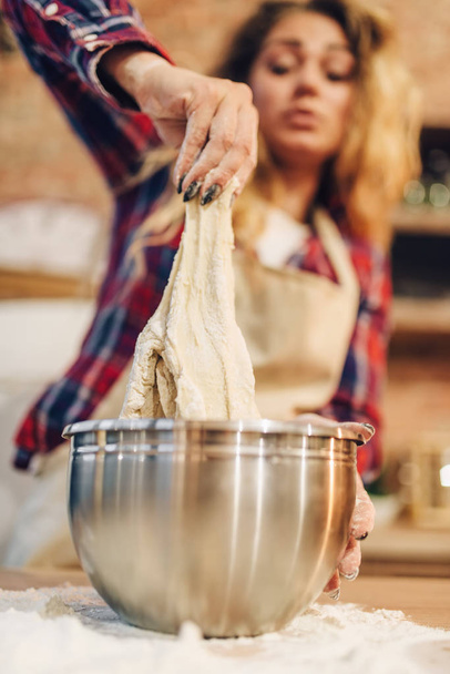 Домохозяйка в фартуке готовит тесто в металлической миске, кухонный интерьер на заднем плане. Женщина-повар готовит свежий домашний торт. Подготовка домашнего пирога
 - Фото, изображение