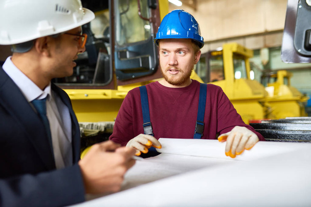 Handsome red-haired worker wearing protective helmet and overall and his superior sharing ideas with each other while studying blueprint at production department of heavy vehicle factory - Φωτογραφία, εικόνα