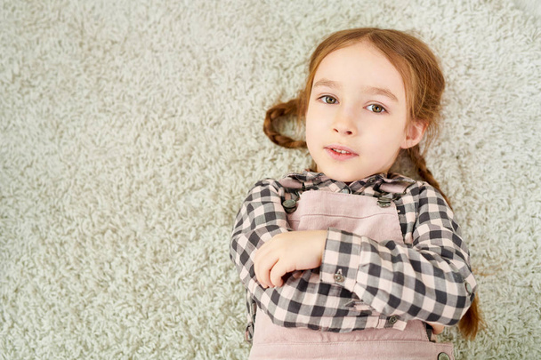 Taille-staand van schattig klein meisje met twee vlechten liggend op gezellige tapijt en camera te kijken - Foto, afbeelding