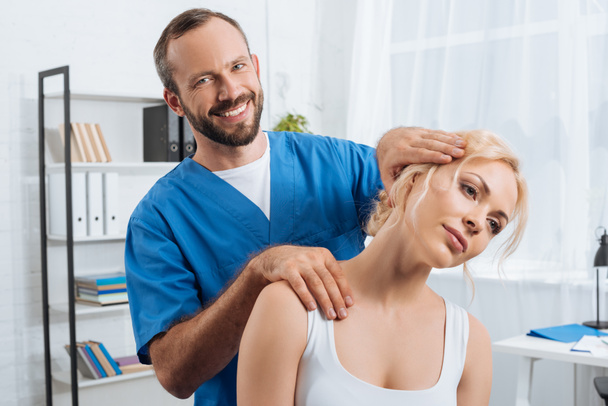 portrait of smiling chiropractor stretching neck of woman during appointment in hospital - Foto, afbeelding