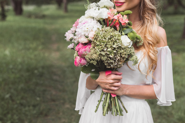 cropped shot of tender smiling blonde bride holding wedding bouquet outdoors - Photo, Image