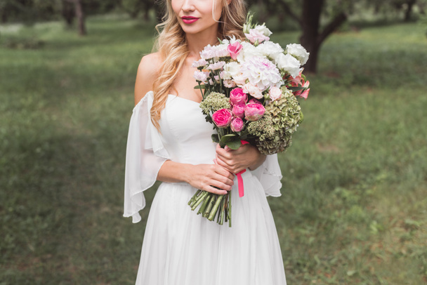 cropped shot of beautiful tender young bride holding wedding bouquet outdoors - Photo, image
