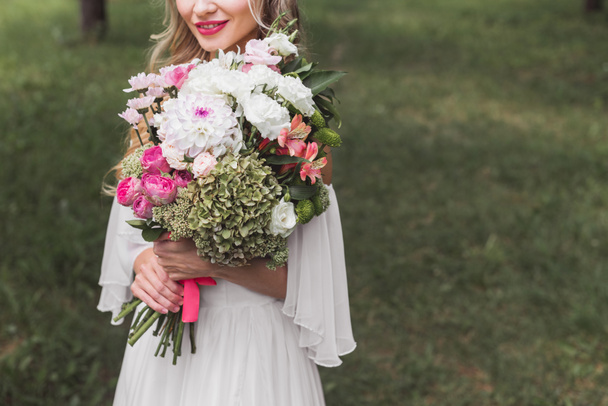 plan recadré de souriant jeune mariée tendre tenant bouquet de mariage en plein air
 - Photo, image