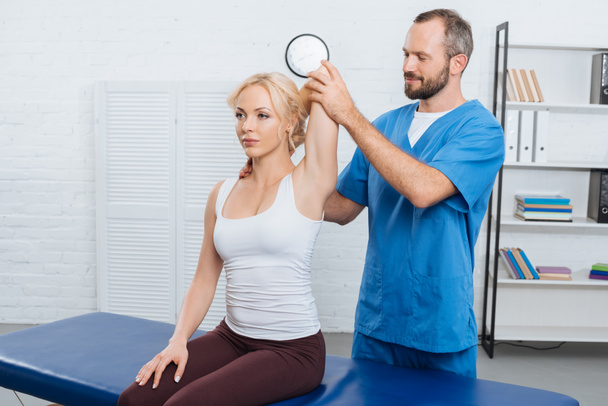 Lächelnder Chiropraktiker streckt Frauenarm auf Massagetisch in Klinik - Foto, Bild