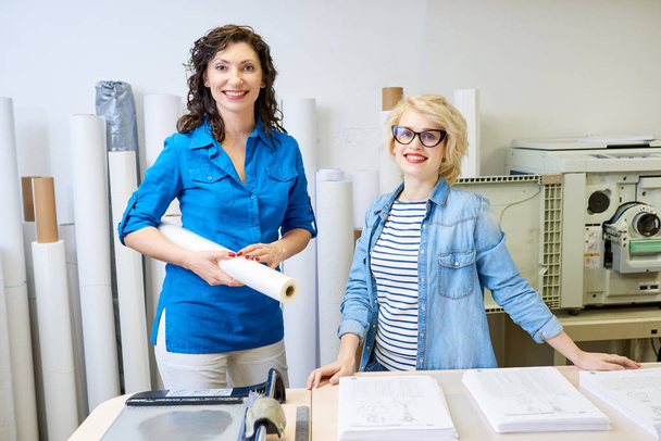 Dos hermosas mujeres sonriendo y mirando a la cámara mientras están de pie en la oficina de impresión juntos
.  - Foto, Imagen