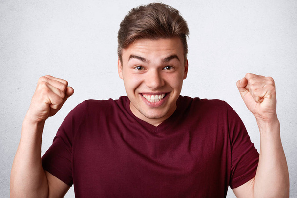 Overjoyed Caucasian male clenches fists as celebrates his success, has broad smile, being in good mood, poses against white concrete wall. Happy positive young man gestures, expresses happiness - Fotoğraf, Görsel