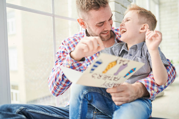 entzückender kleiner Junge und sein bärtiger Papa amüsieren sich zusammen beim Vorlesen einer handgefertigten Grußkarte für den Vatertag, Panoramafenster im Hintergrund - Foto, Bild