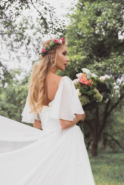 beautiful blonde bride in wedding dress and floral wreath holding bouquet of flowers in park - Φωτογραφία, εικόνα