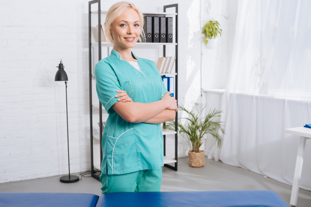 portrait of smiling female masseur with arms crossed standing at massage table in clinic - Photo, Image