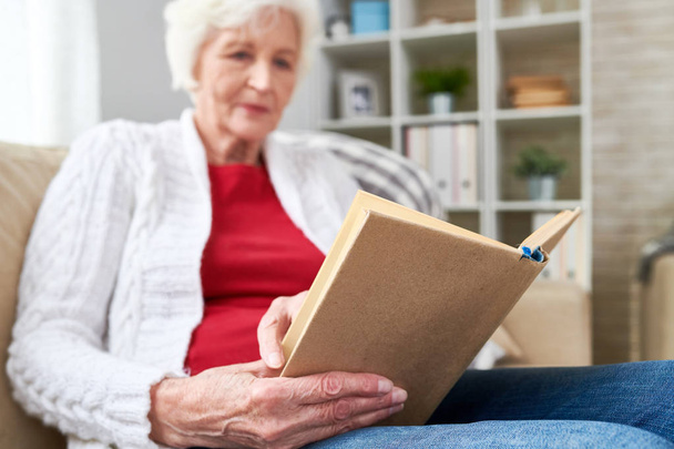 Low angle portrait of white-haired woman reading book while enjoying leisure time at home, focus on book in wrinkled hands - Foto, imagen