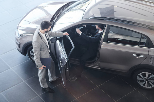 Высокий угол обзора в полный рост у продавца, открывающего дверь для клиента, который садится в свой новенький роскошный автомобиль
 - Фото, изображение