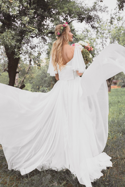 back view of beautiful blonde bride in wedding dress holding bouquet of flowers and walking in park - Foto, Imagem
