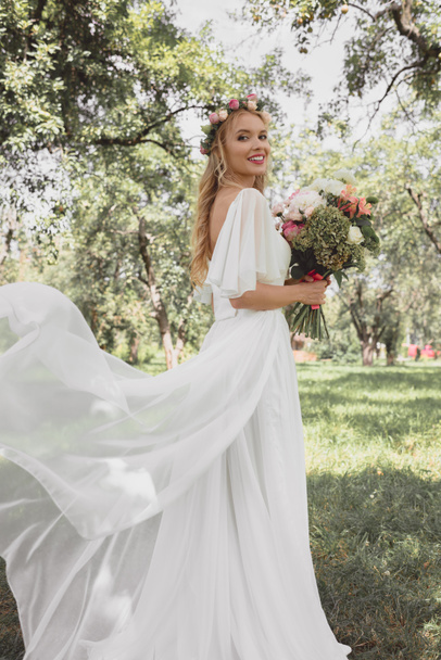 beautiful young bride in wedding dress holding bouquet of flowers and smiling at camera in park - Fotografie, Obrázek
