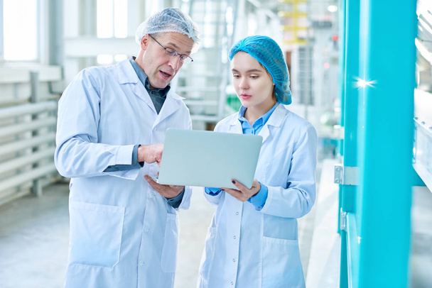 Portrait of senior factory worker wearing lab coat training young woman and using laptop while standing in clean production workshop of modern plant - Foto, Bild