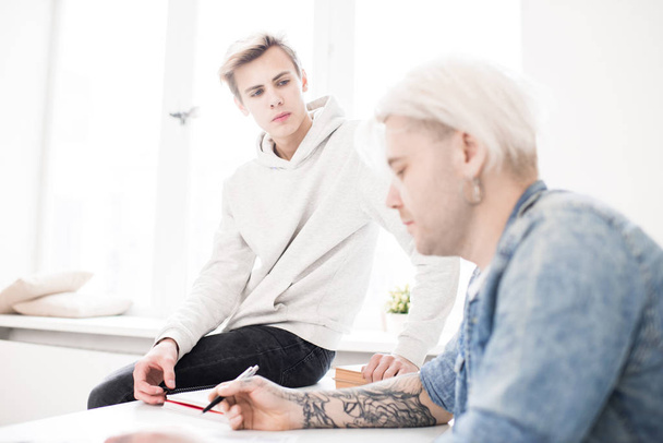 Young Caucasian male student sitting on desk and listening attentively to his adult college mate with tattooed hands - Foto, Imagem