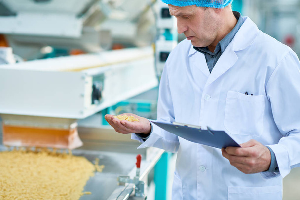Mid section portrait of senior factory worker checking production process standing by conveyor belt holding handful of dry macaroni, copy space - Photo, Image