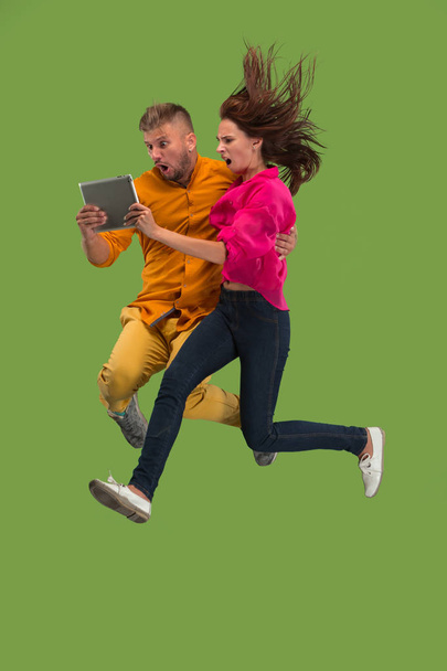 Jump of young couple over green studio background using laptop or tablet gadget while jumping. - Fotografie, Obrázek