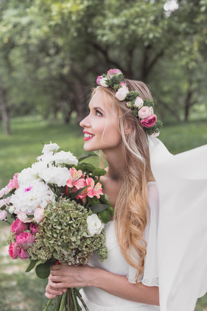 vista lateral de hermosa novia joven sonriente en velo celebración de ramo de boda y mirando hacia arriba
 - Foto, Imagen